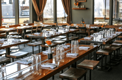 A spacious restaurant interior with wooden tables set for dining, featuring flowers and glassware on each table.