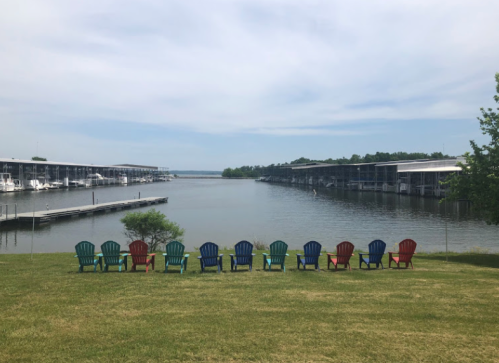Colorful Adirondack chairs line a grassy area overlooking a calm marina with boats and a cloudy sky.