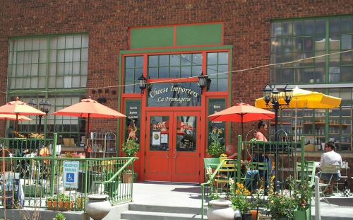 A charming cheese shop with outdoor seating, colorful umbrellas, and a brick facade, inviting customers to enjoy the ambiance.