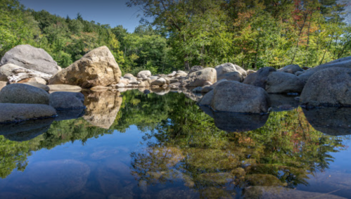 A serene river scene with smooth rocks and trees reflecting in calm water under a clear blue sky.