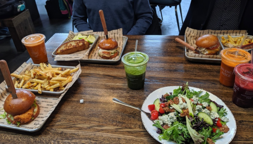 A table with two trays of burgers and fries, and a colorful salad with drinks in jars.