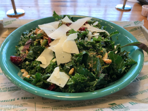 A vibrant salad with kale, cranberries, pine nuts, and shaved cheese in a teal bowl on a tray.