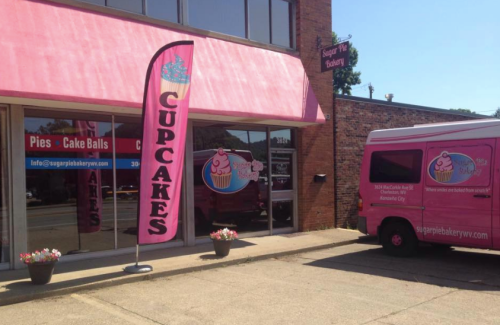 A pink bakery storefront with a cupcake sign and a matching delivery van parked outside.
