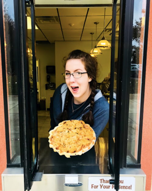 A young woman with glasses smiles while holding a pie at a restaurant window.