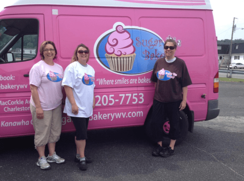 Three women stand in front of a pink food truck with a cupcake logo, wearing matching t-shirts and smiling.
