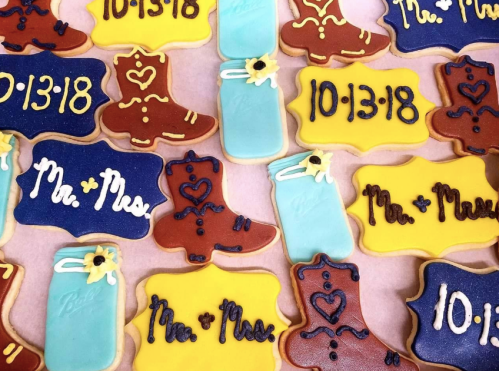 Colorful decorated cookies featuring cowboy boots, wedding dates, and "Mr. & Mrs." designs on a pink background.