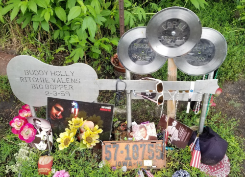 Memorial for Buddy Holly, Ritchie Valens, and Big Bopper with metal plaques, flowers, and photos at a gravesite.