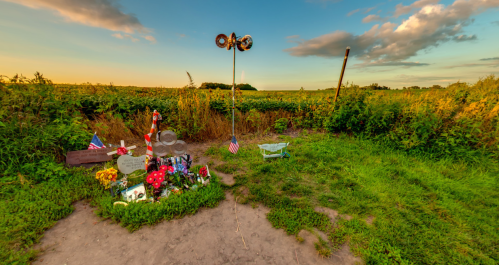 A small memorial in a grassy field, adorned with flowers, flags, and personal items, under a colorful sky.