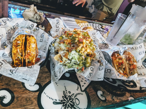 Three colorful tacos and a bowl of nachos on a wooden table, wrapped in branded paper.