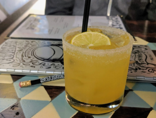 A cocktail with a salted rim and lemon slice sits on a colorful table, with a menu and pencil in the background.
