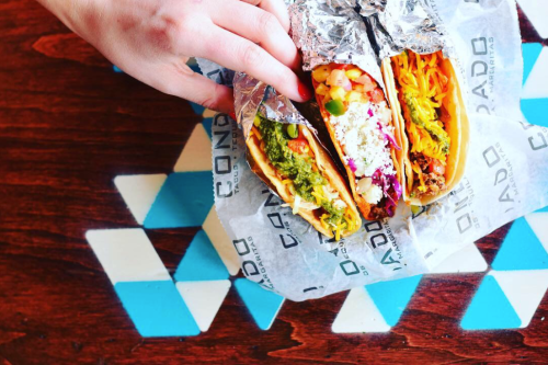 A hand holding three colorful tacos wrapped in foil, placed on a patterned wooden table.