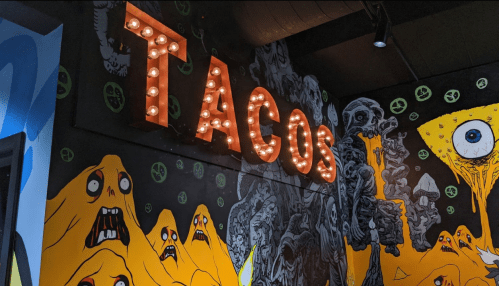 A vibrant mural featuring spooky characters with a lit "TACOS" sign in a colorful, artistic setting.