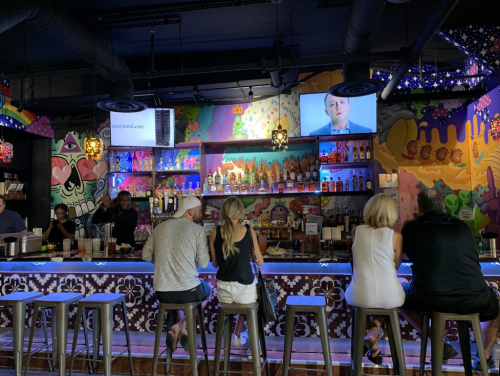 A vibrant bar scene with colorful murals, patrons sitting at the counter, and a TV displaying a presentation.