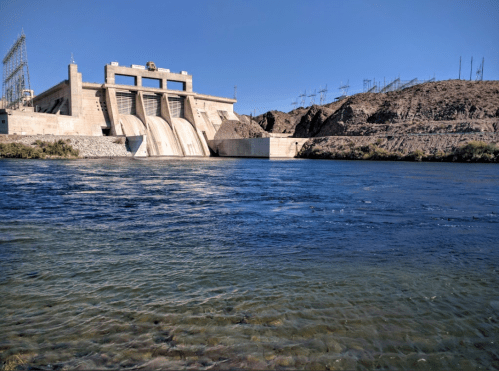 A large dam with flowing water, surrounded by rocky terrain and power lines under a clear blue sky.