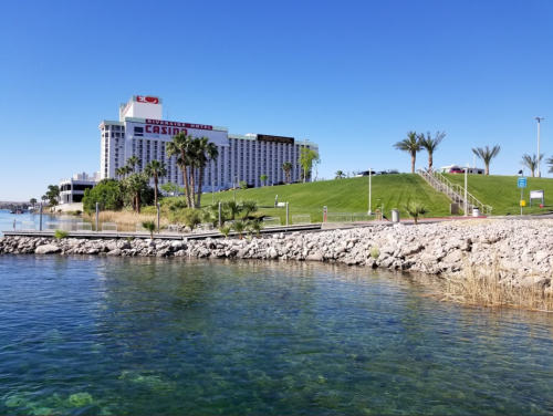 A clear view of a casino building beside a calm lake, with palm trees and a grassy hill in the background.