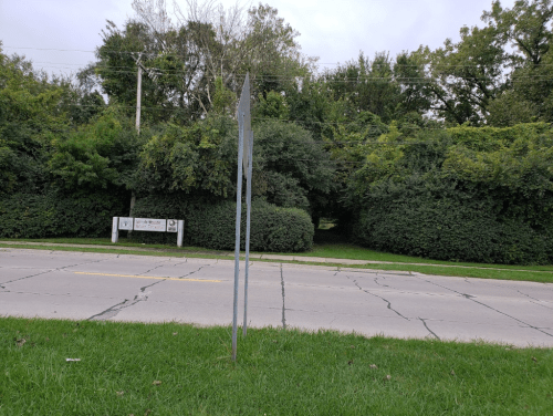 A street sign stands beside a cracked road, with dense greenery and a path visible in the background.