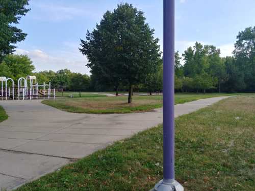 A park scene featuring a tree, grassy areas, and a winding path near playground equipment in the background.