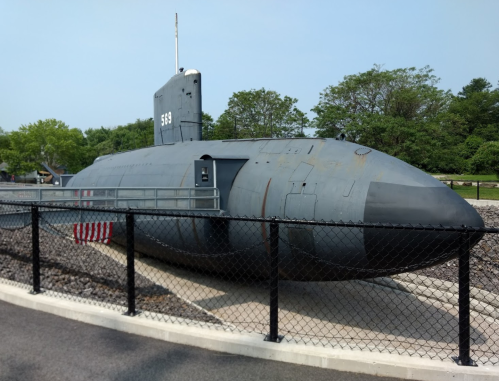 A gray submarine displayed outdoors, surrounded by a fence and greenery, with an American flag visible nearby.