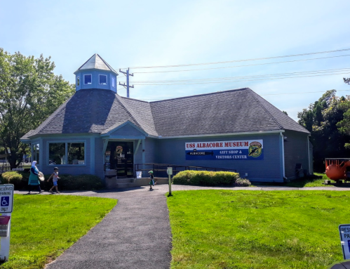 Exterior of the USS Albacore Museum, a blue building with a sign, surrounded by grass and visitors.