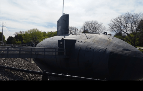 A large black submarine displayed on land, with a walkway leading to its entrance and trees in the background.