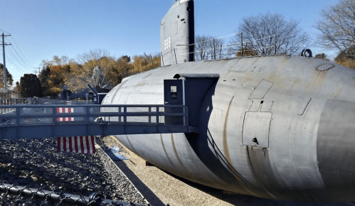 A large submarine on display, with a walkway leading to its entrance, surrounded by trees and a fence.