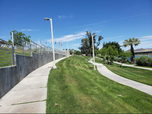 A winding pathway through a green park with palm trees and flags in the background under a clear blue sky.