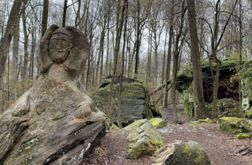 A stone sculpture resembling a figure sits among trees and rocks in a wooded area, with a path leading further into the forest.