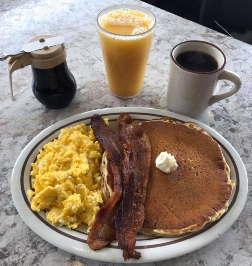 A breakfast plate with pancakes, bacon, scrambled eggs, and a side of syrup, accompanied by orange juice and coffee.