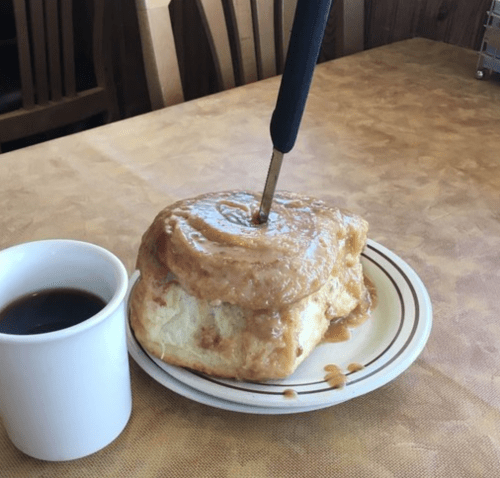 A large cinnamon roll with a knife inserted, next to a cup of coffee on a table.