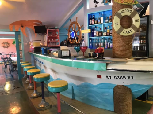 Colorful bar with a boat design, palm tree decor, and stools, set in a vibrant lakeside-themed interior.