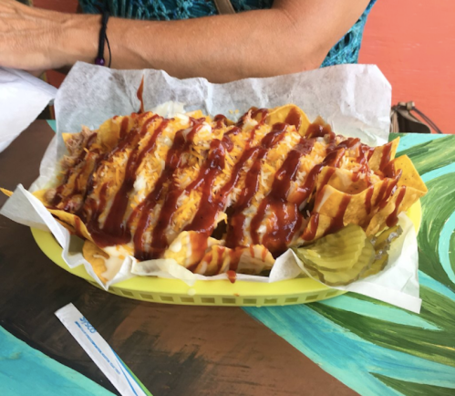 A vibrant plate of nachos topped with cheese, sauce, and pickles, served in a yellow basket on a colorful table.