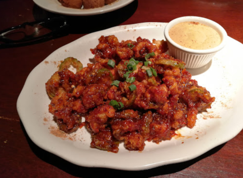 A plate of crispy fried Brussels sprouts drizzled in a spicy sauce, served with a small cup of dipping sauce.