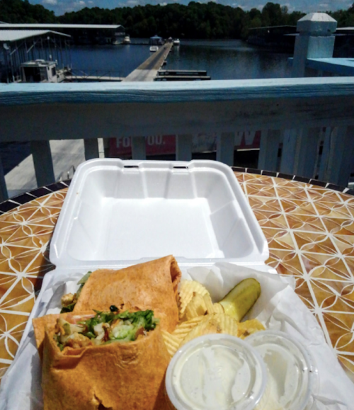 A wrap with lettuce and other fillings, served with chips and pickles, overlooking a marina and water.