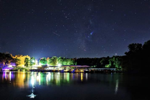 A serene night scene of a lake reflecting colorful lights and stars, surrounded by trees and a dock.