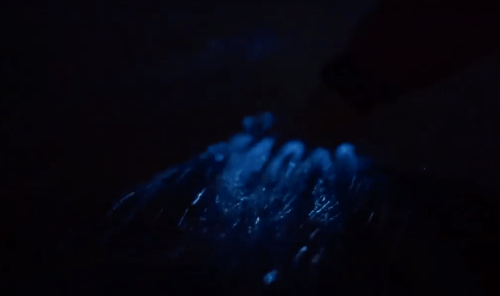 A hand reaching into water, creating ripples illuminated by a blue light in a dark setting.