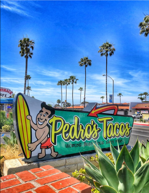 Colorful sign for Pedro's Tacos featuring a surfer, palm trees, and a bright blue sky.