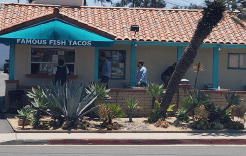 A colorful taco stand with a tiled roof, surrounded by desert plants and people ordering food.