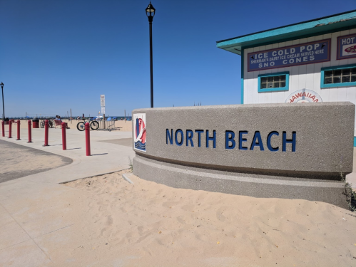 Sign for North Beach with sandy path and beach amenities in the background under a clear blue sky.