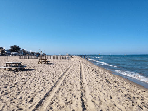A sandy beach with picnic tables, gentle waves, and a clear blue sky, leading to a pier in the distance.