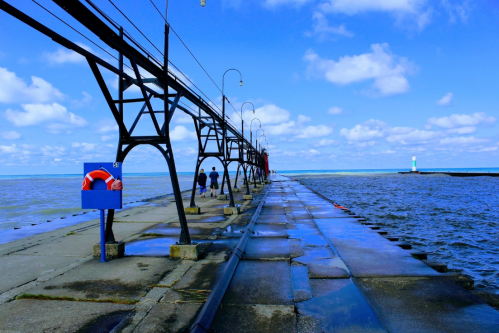 A long pier extends over water, with people walking and a lighthouse visible in the distance under a blue sky.