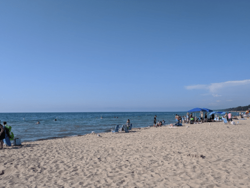 A sunny beach scene with people swimming, lounging on sand, and umbrellas near the water under a clear blue sky.