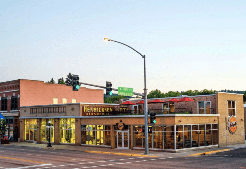 A modern restaurant with outdoor seating, located at a street intersection, featuring large windows and a vibrant sign.