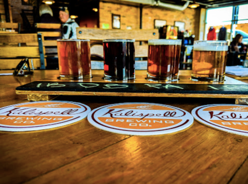 A flight of four craft beers on a wooden paddle, with Kalispell Brewing Co. coasters on a rustic table.