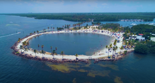 Aerial view of a circular beach surrounded by palm trees, with people swimming and relaxing in the water.