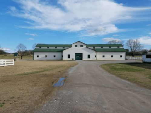 A large white barn with a green roof, surrounded by a dirt path and open fields under a blue sky with clouds.