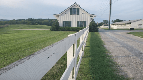 A white farmhouse with green landscaping, viewed from a gravel driveway lined with a white fence.