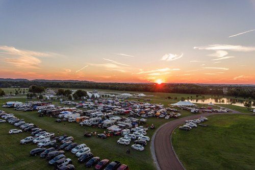 Aerial view of a bustling outdoor event at sunset, with rows of cars and tents set up in a grassy area.