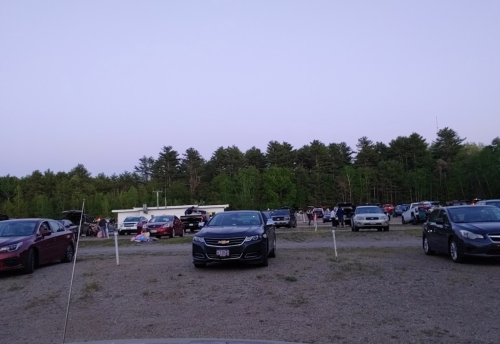 A crowded parking lot with various cars, surrounded by trees and a building in the background during twilight.