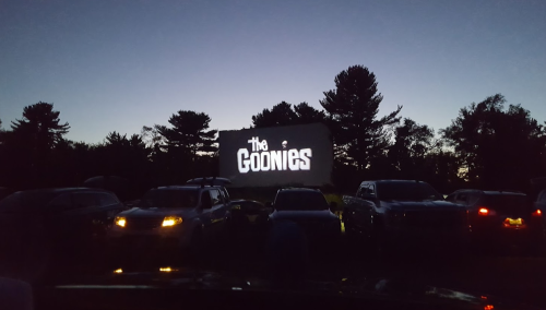 A drive-in movie screen displaying "The Goonies" at dusk, surrounded by parked cars and trees.
