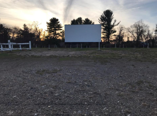 An empty outdoor movie screen at sunset, surrounded by trees and a gravel lot.
